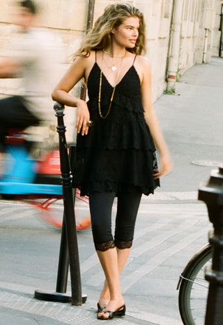 a-woman-stands-on-a-city-sidewalk-wearing-a-black-sleeveless-ruffled-mini-dress-with-thin-straps-black-capri-leggings-with-lace-trim-and-black-flat-sandals-she-is-accessorized-with-layered-gold-necklaces