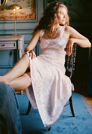 a-woman-reclining-on-a-dark-wooden-chair-in-a-vintage-style-room-wearing-a-pale-pink-lace-trimmed-maxi-dress-and-layered-gold-necklaces
