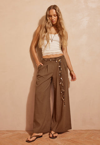 a-woman-stands-against-a-textured-beige-wall-wearing-wide-leg-brown-pants-with-a-decorative-hanging-chain-detail-at-the-side-a-white-cropped-tank-top-and-black-sandals